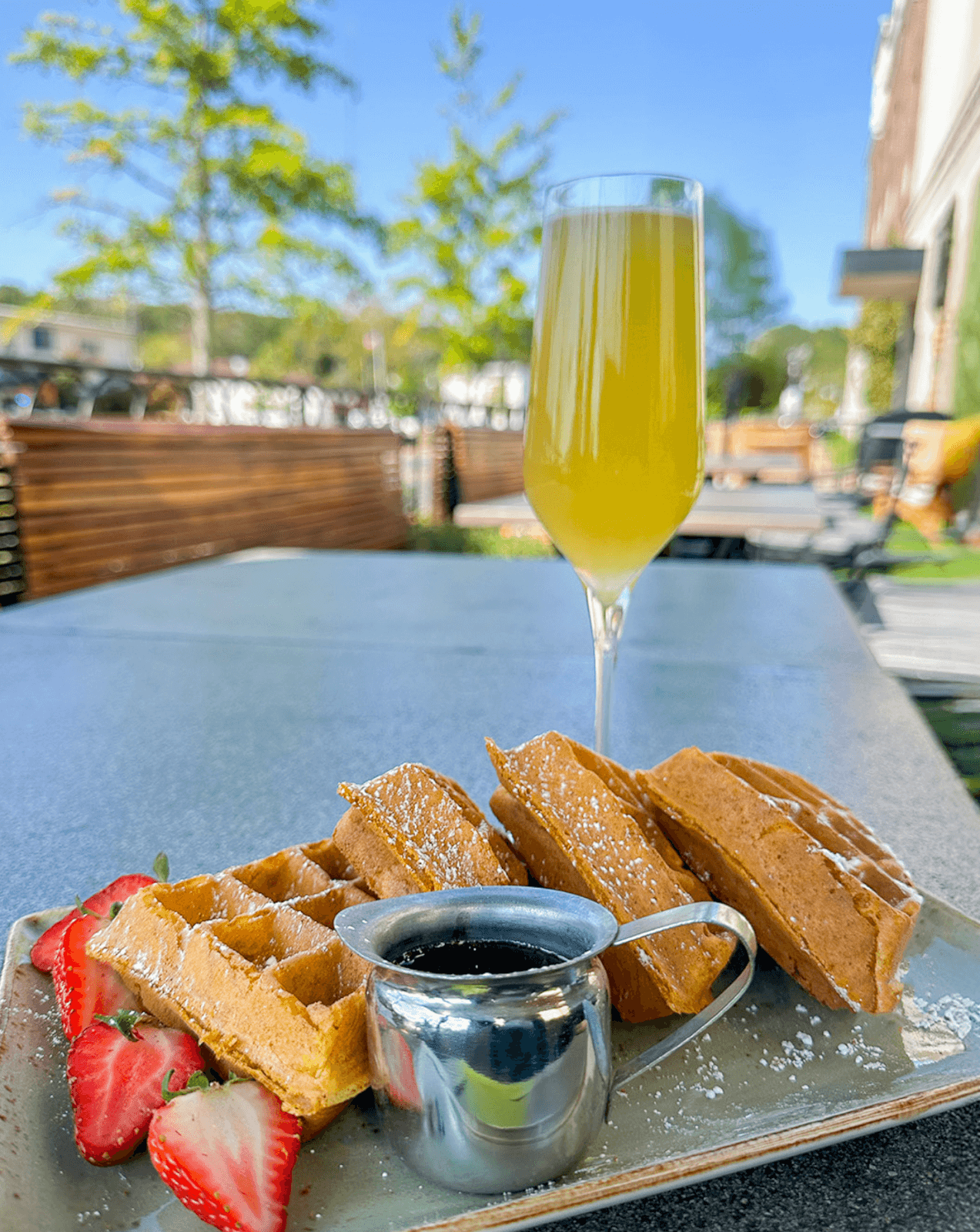 Waffles dusted with powdered sugar, fresh strawberries, and syrup in a small pitcher sit on the outdoor table of a cozy BHAM cafe, accompanied by a glass of mimosa. 