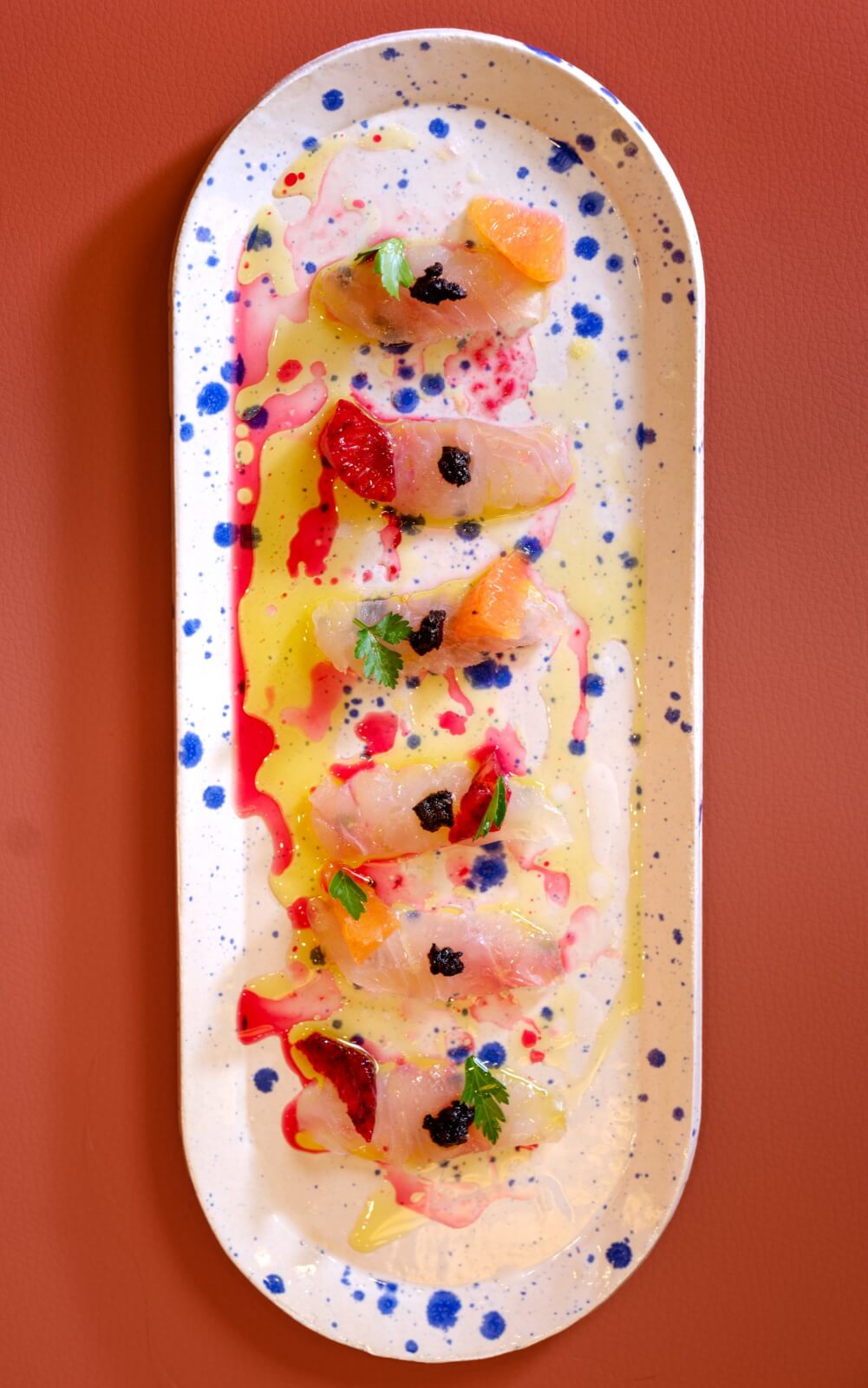 An oval plate with five slices of raw fish adorned with herbs, black roe, and colorful sauces graces the terracotta surface. 