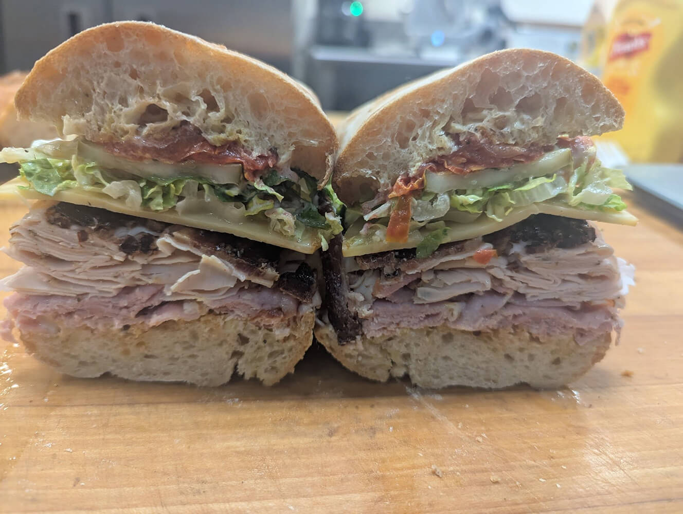 A beautifully crafted sandwich featuring layers of turkey, ham, lettuce, tomato, and cheese nestled within ciabatta bread sits elegantly on a wooden surface—a delightful concept inspired by the vibrant culinary scene at new restaurants in Birmingham.