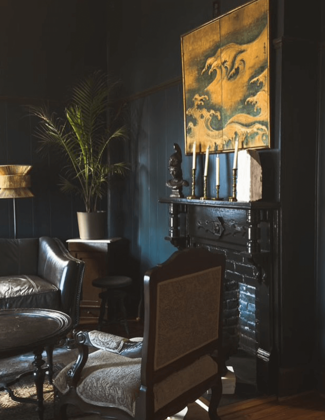 In a dimly lit room with dark blue walls and a black fireplace, beneath a painting, the ambiance evokes vintage charm. A leather sofa, upholstered chairs, and potted plant enhance the cozy concept.