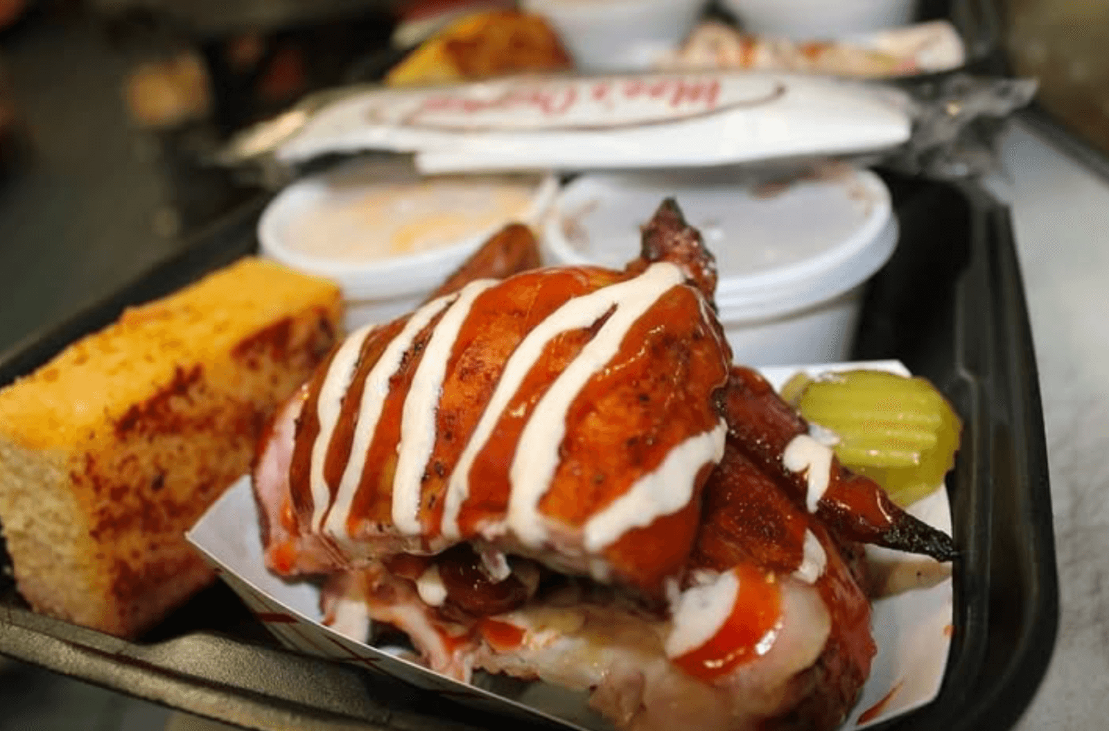 BBQ chicken slathered in sauce with a white drizzle, served alongside cornbread and pickles, all presented on a tray with small containers of sides.