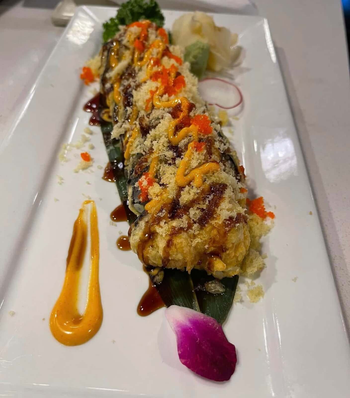 A sushi roll topped with sauces and vibrant orange roe rests elegantly on a rectangular white plate, garnished with a radish slice and a delicate purple flower petal. Discover this culinary delight at one of Birmingham’s new restaurants to try this month.