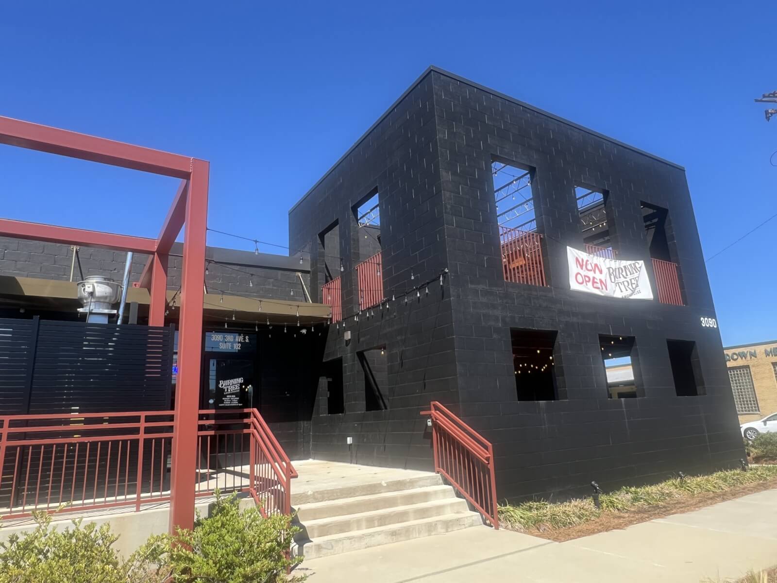 Black two-story building in Birmingham with a "Now Open" sign on the upper level, featuring red accents and stairs leading to the entrance. New restaurant concepts emerge under the clear blue sky.