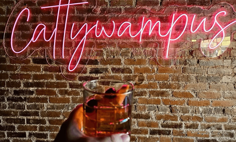 A hand holds a cocktail with fruit garnish in front of a brick wall featuring a neon sign that reads "Cattywampus," one of Birmingham's exciting new dining concepts.
