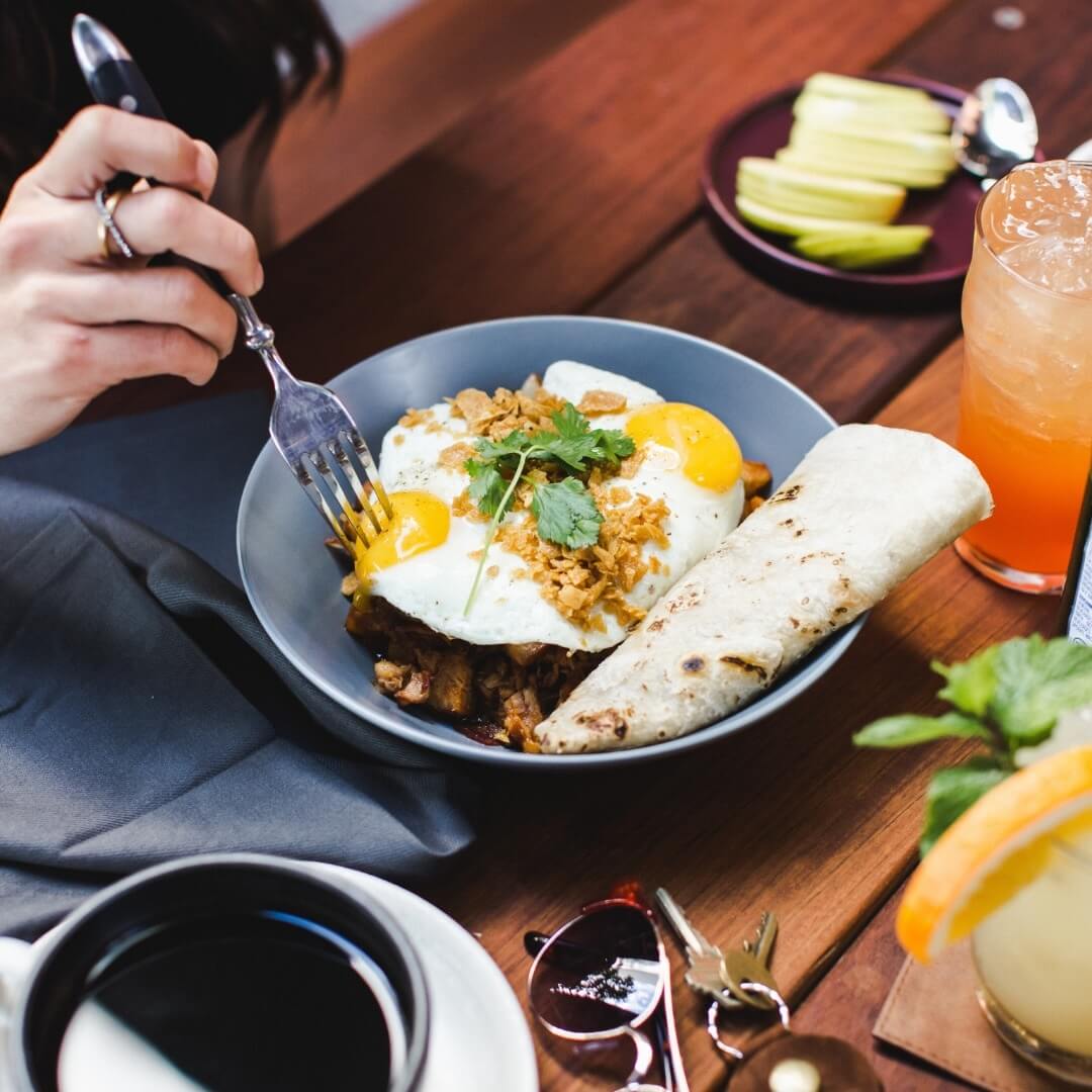 A person enjoys Easter Brunch, holding a fork over a plate with fried eggs, cilantro, and a rolled tortilla. Nearby sit coffee, sunglasses, and apple slices on the wooden table—a serene BHAM morning scene.