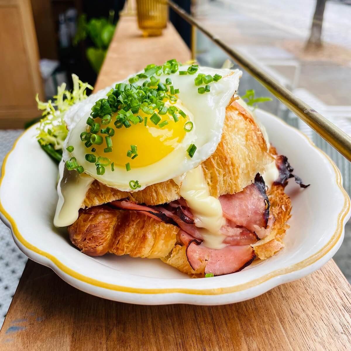 This delightful croissant sandwich, perfect for an Easter brunch, features ham, melted cheese, and a sunny-side-up egg sprinkled with fresh chives. Served on a white plate alongside crisp greens, it's a sumptuous BHAM treat to savor.