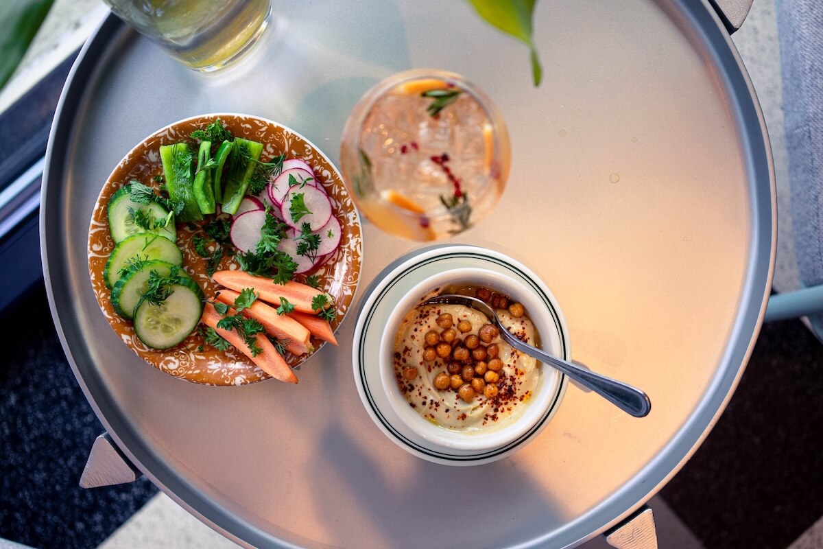 A round table set for Easter Brunch features a bowl of creamy dip topped with chickpeas alongside a plate of sliced vegetables, like cucumber, radish, and carrots. Above, a refreshing drink garnished with herbs adds an elegant touch to the Bham gathering.