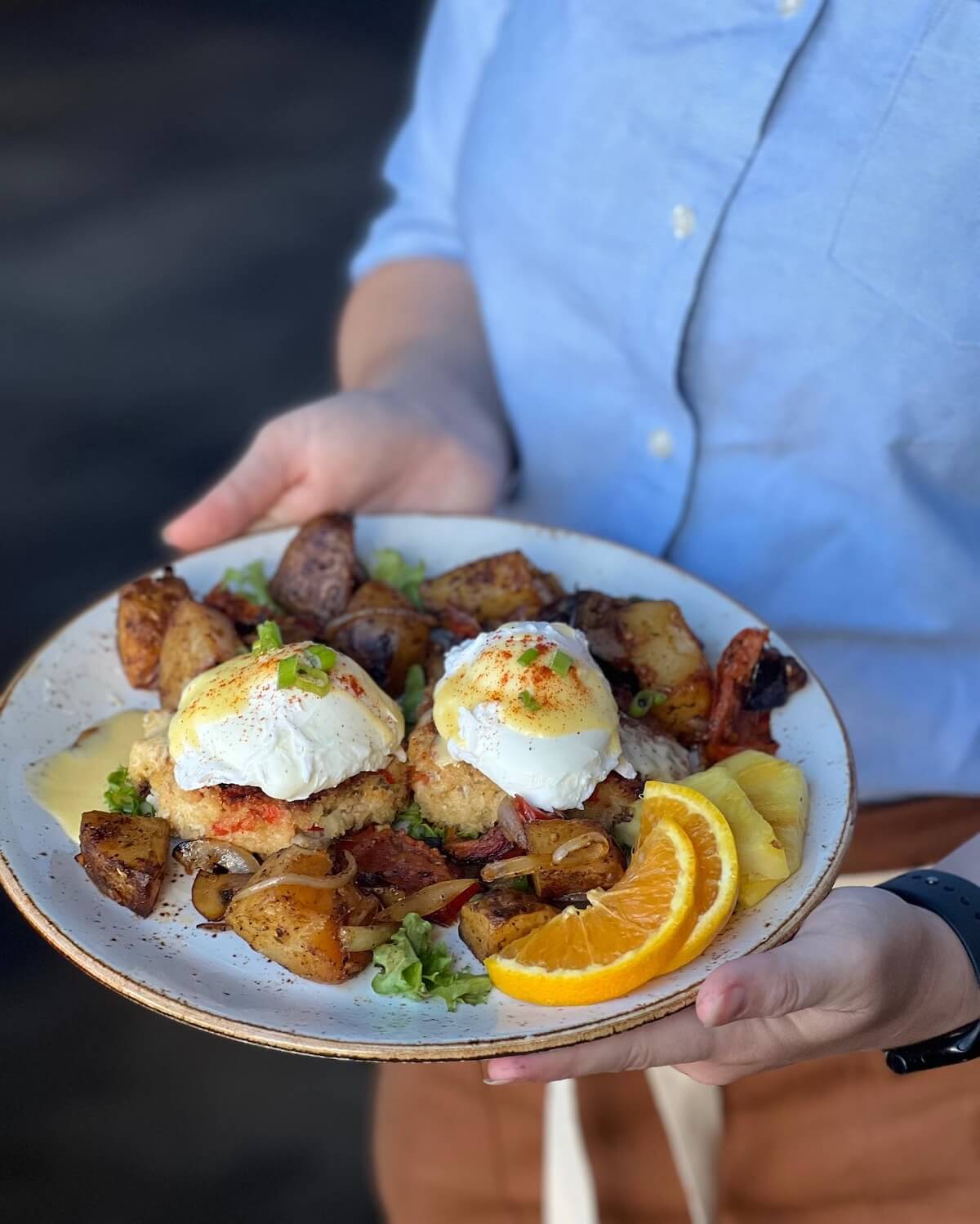 A person holds a plate of poached eggs on crab cakes, surrounded by roasted potatoes and orange slices, all garnished with green onions—perfect for an elegant Easter Brunch in BHAM.