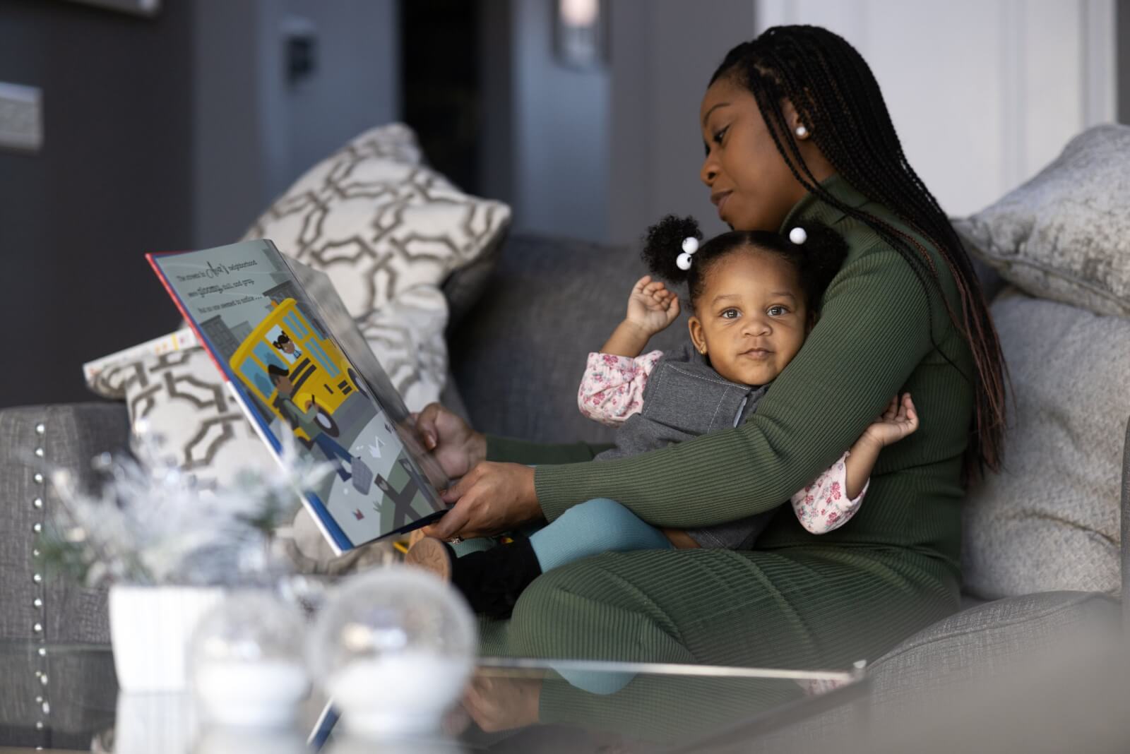 A mother sits with her young daughter, around 2 years old, in her lap and reads a book to her. 