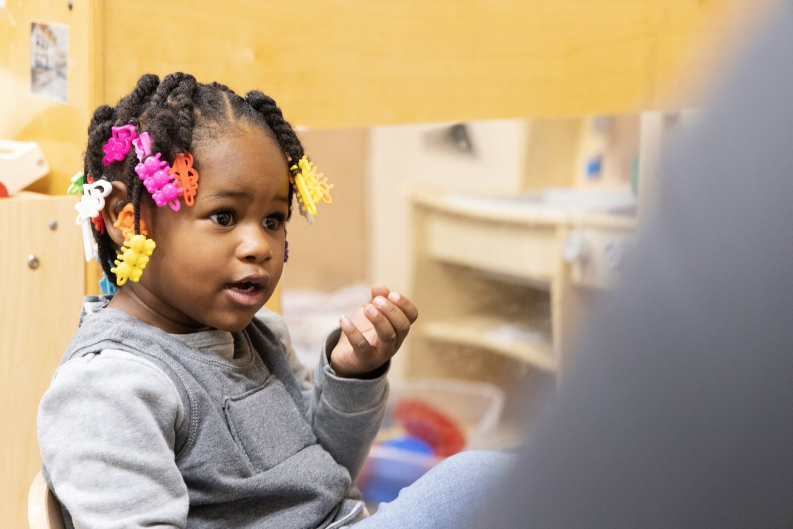 A young girl, around 2 or 3 years old, with brightly colored clips in her hair looks at someone beyond the camera and appears to be talking to them.