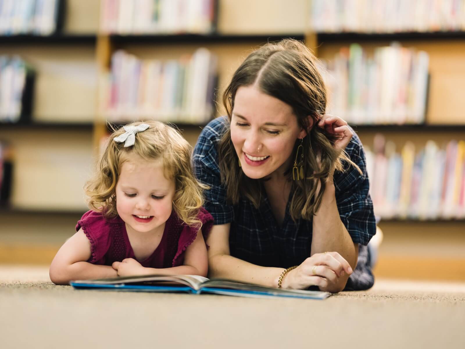 Ruth Ann Moss reads a book next to a young girl, around 3 years old, with a bookcase blurred in the background.