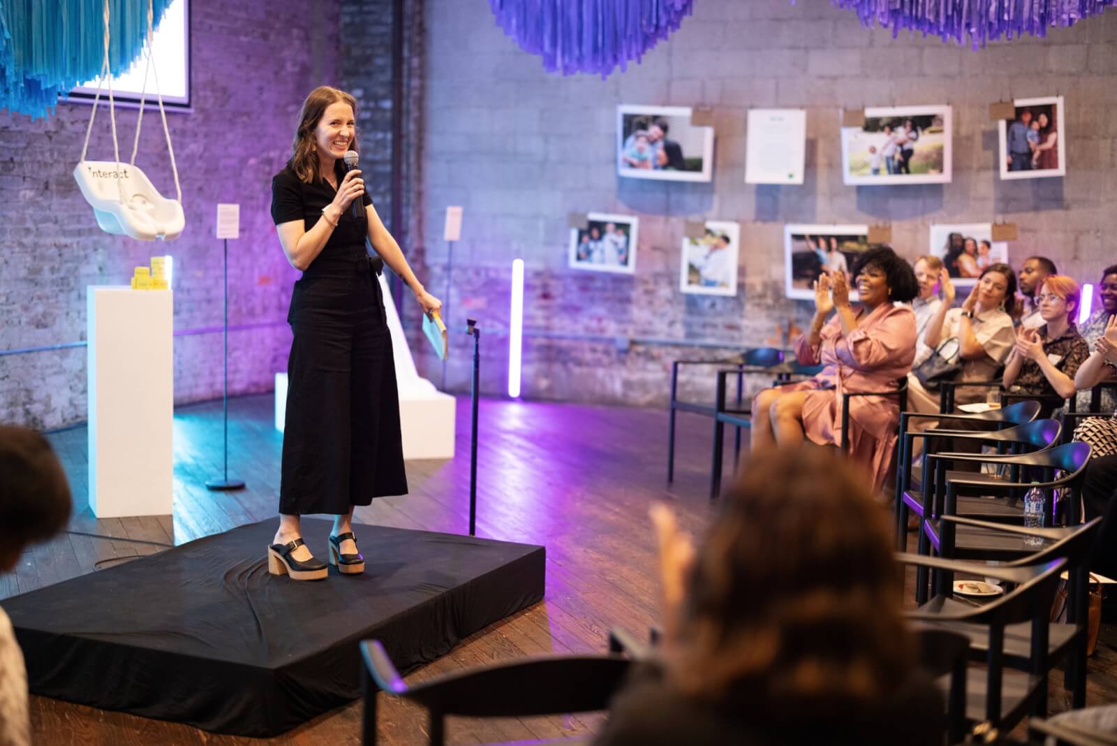 Ruth Ann Moss wears a black dress and speaks into a microphone on a small stage. The audience, seated and clapping, is surrounded by photos and a swing seat on display at a Small Magic event.