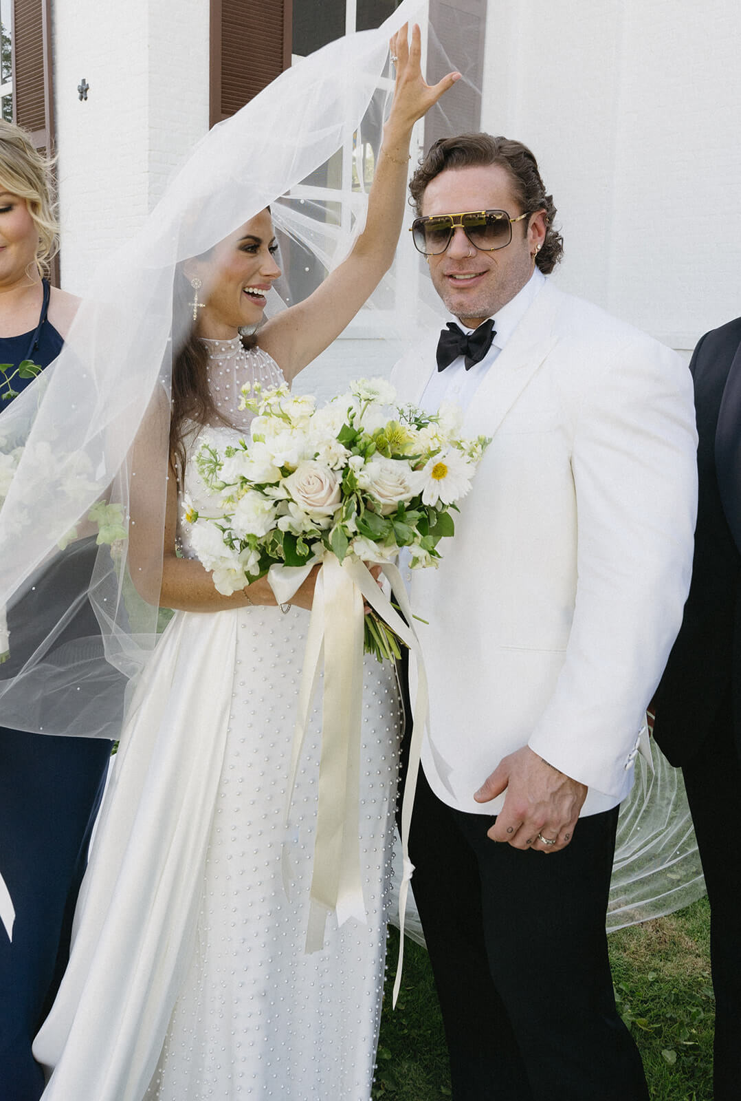 Bride in white dress with a floral bouquet holds up her veil next to a man in a white jacket and sunglasses, both smiling radiantly outside a historic Pensacola wedding venue.