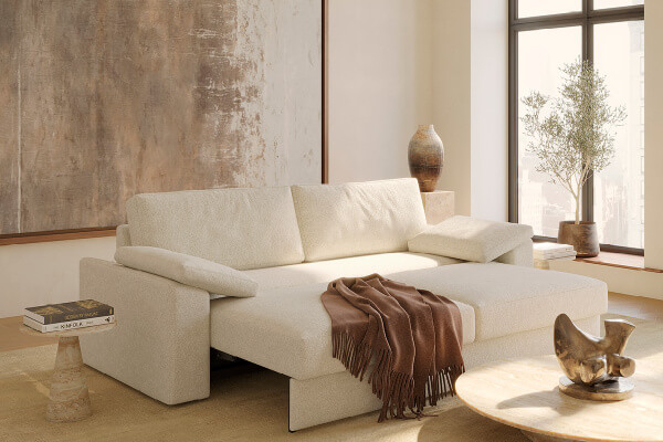 A beige sofa with a brown throw blanket sits in a sunlit room, embodying Scandinavian design. Nearby, a round wooden table holds books and potted plants, while a decorative sculpture rests on a stack of reads.