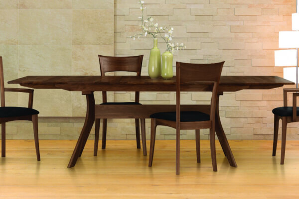 A wooden dining table with four chairs sits on a light wood floor against a stone wall, embodying Scandinavian design. Two green vases with branches grace the table, creating an inviting and minimalist vibe.