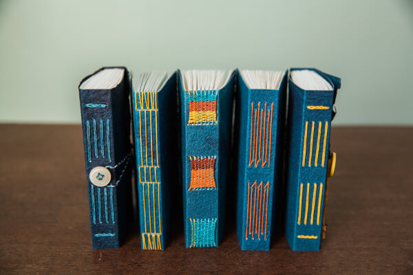 Five handmade books with unique stitch patterns and colorful spines are displayed upright on a wooden surface, showcasing variations of long stitch bookbinding techniques.