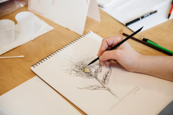 Amid a workshop series, someone paints a botanical illustration of a flower in a sketchbook on a wooden table, surrounded by art supplies.