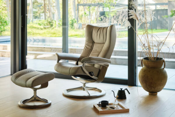 A tan recliner and matching footrest, showcasing elegant Scandinavian design, are near a large window. A wooden tray with cups and a teapot rests on the floor. A potted plant completes the scene, offering a tranquil view of the patio and greenery outside.