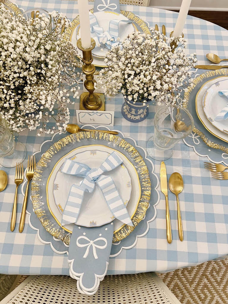 Elegant table setting by SJ Bailey featuring a blue checkered tablecloth, gold utensils, white plates adorned with blue bows, and exquisite floral centerpieces.