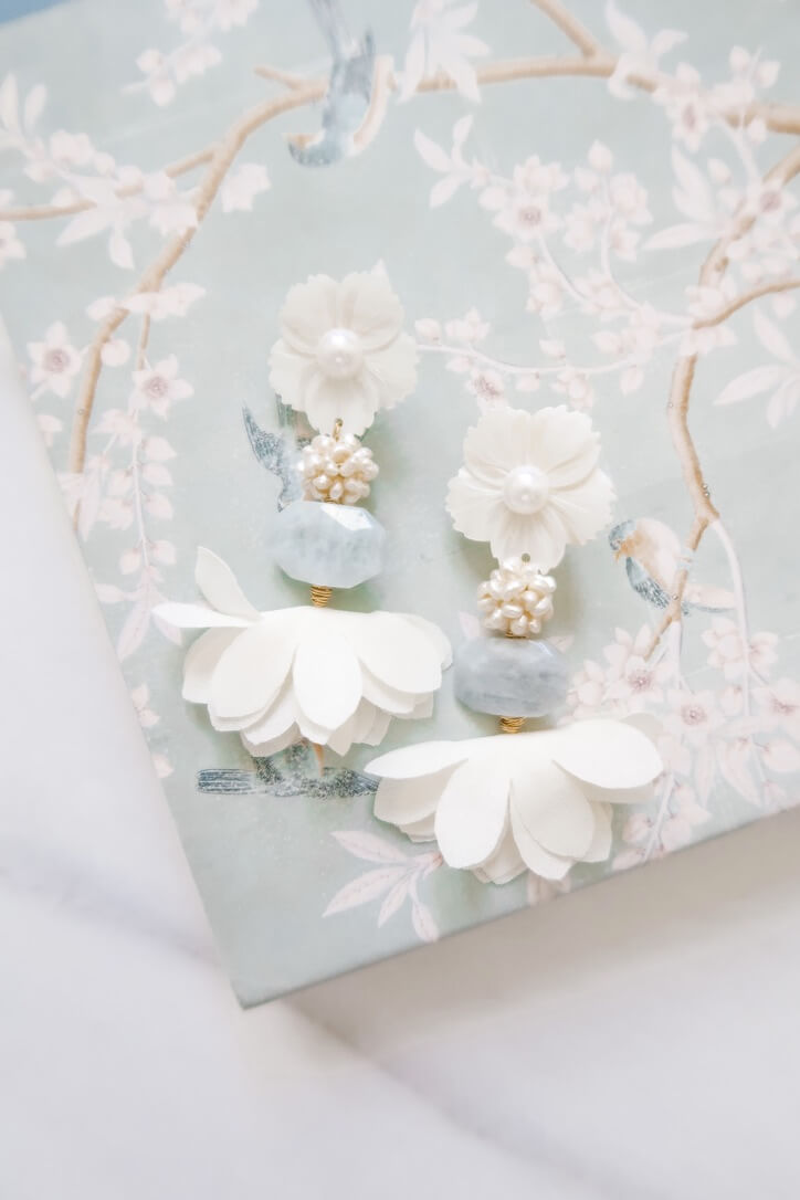 SJ Bailey's floral-themed earrings, adorned with elegant white and pearl accents, rest gracefully on a floral-patterned surface in soft pastel hues, capturing a timeless sense of beauty.