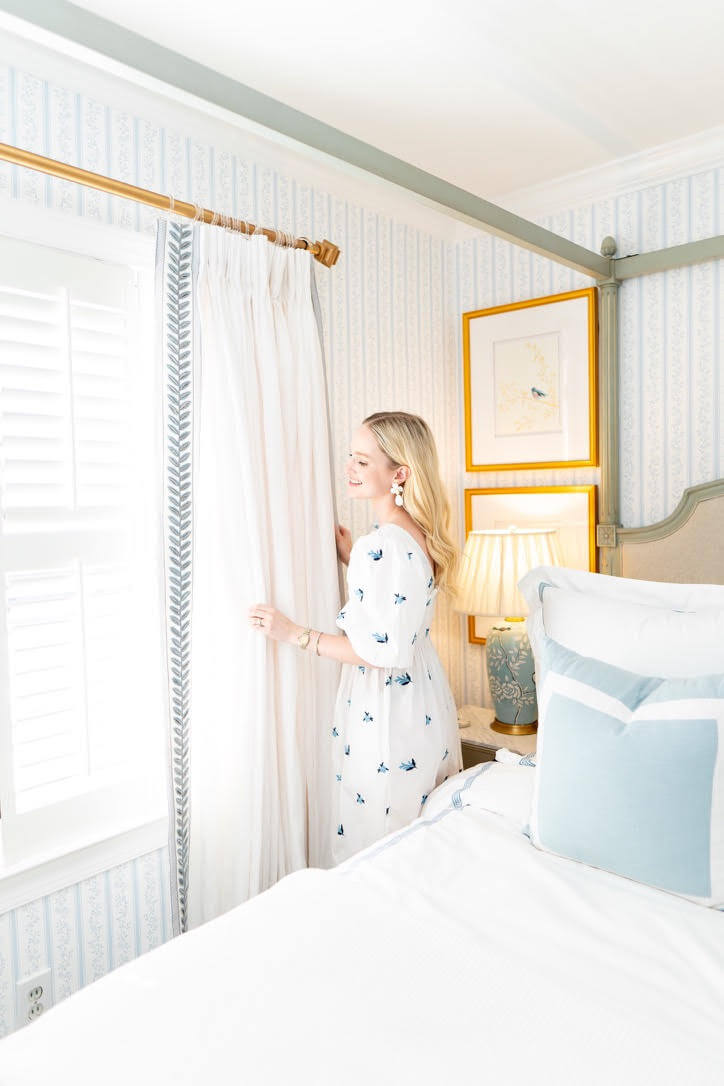 In a bright, well-lit room adorned with pastel decor, a person in a white dress adjusts the curtain beside a bed.