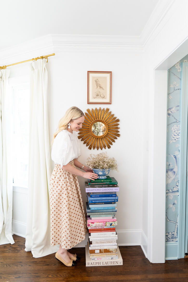 A person stands beside a tall stack of books topped with flowers in a bright room. The wall features a circular sunburst mirror and framed art, echoing the timeless elegance of an SJ Bailey design.