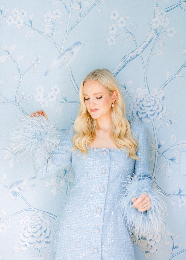 A woman in a light blue, textured dress with feathered sleeves stands gracefully against a matching floral and bird-patterned wallpaper.