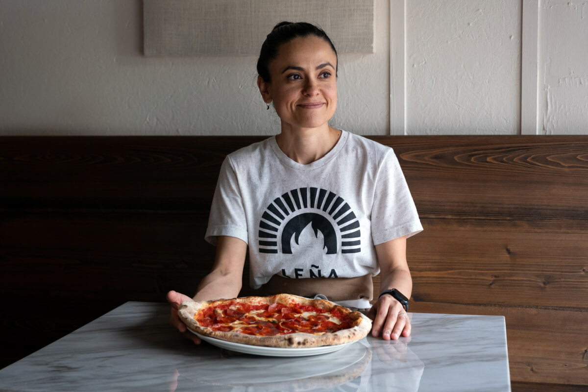 A person sits at a marble table, savoring a Leña Pizza, dressed in a white t-shirt with a black emblem, against the rustic charm of a wooden wall.