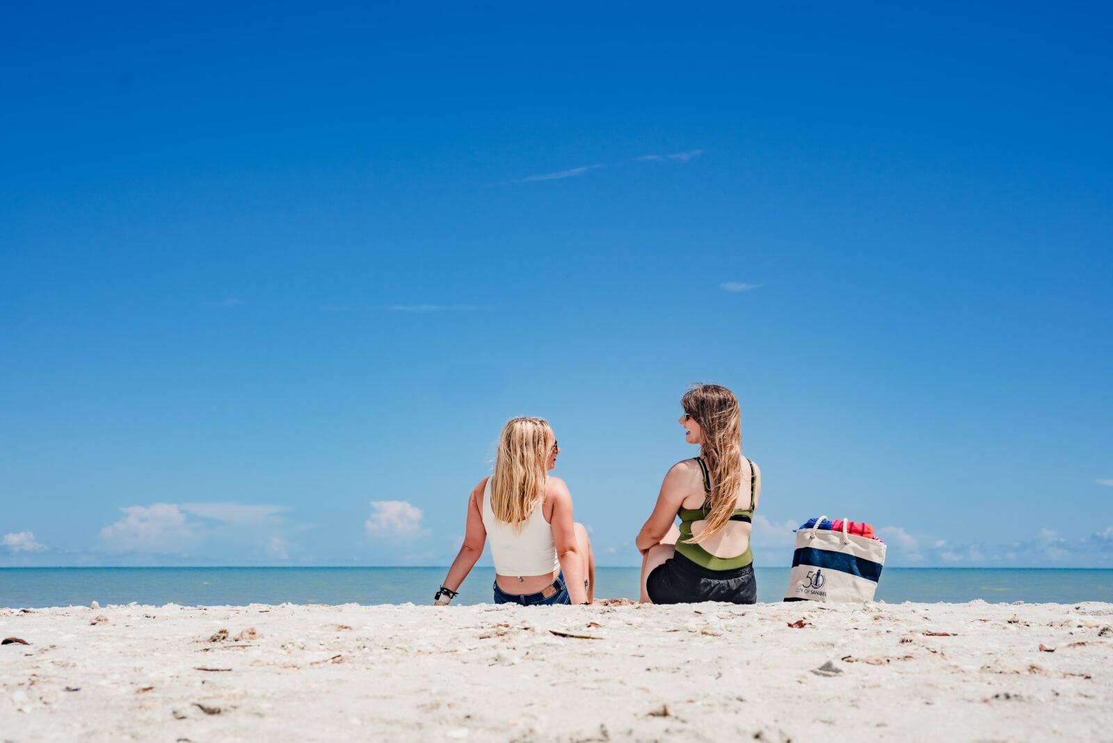 Two people sit on a sandy beach in a tranquil Florida town, facing the ocean under a clear blue sky. A striped bag is placed beside them, a testament to the area's vibrant recovery post-disaster.