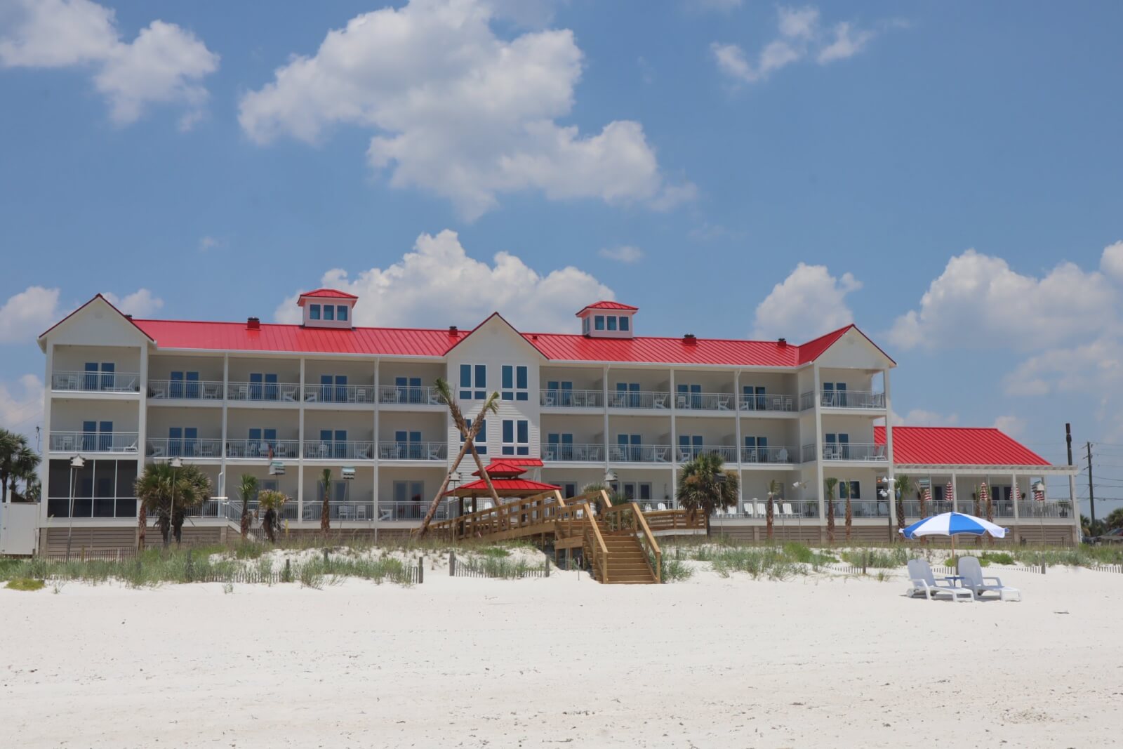 Beachfront hotel with red roofs and multiple balconies sits on a sandy beach under a blue sky. Rebuilt post-disaster, this gem in one of the charming Florida towns offers two beach chairs and an umbrella waiting for you on the sand.