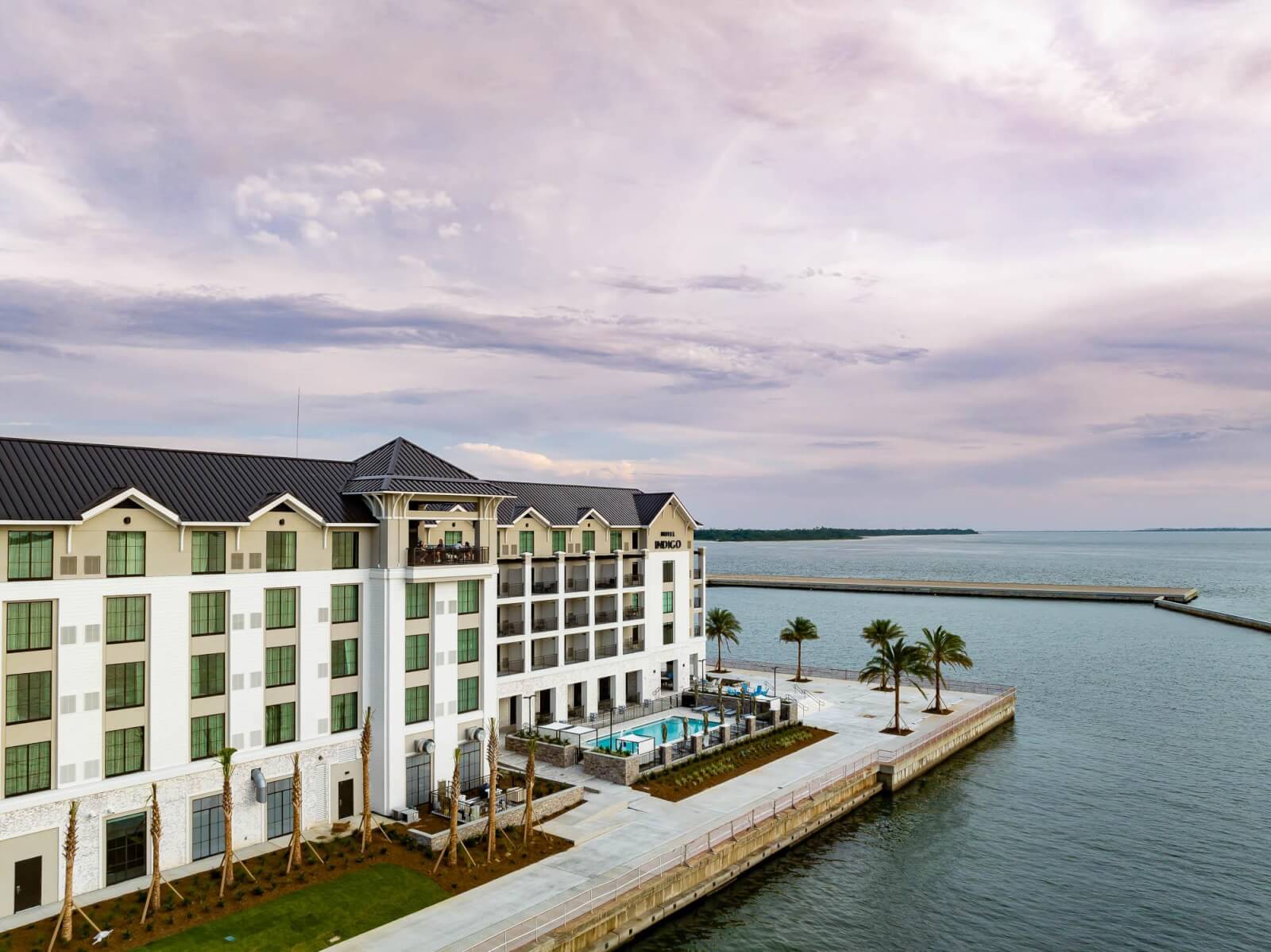 A waterfront hotel rises gracefully over a serene body of water. Rebuilt to perfection post-disaster, it boasts multiple floors with views of palm trees and a sparkling swimming pool beneath the cloudy sky.