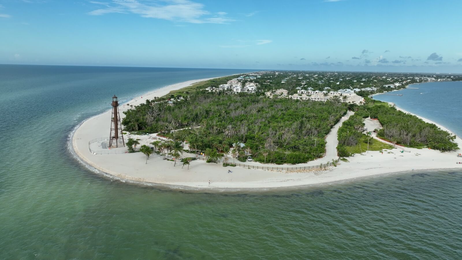 Aerial view of a sandy island featuring a lighthouse surrounded by blue-green water. Once a Florida town, its lush greenery marks the area now rebuilt, with resilient buildings standing proudly in the background.