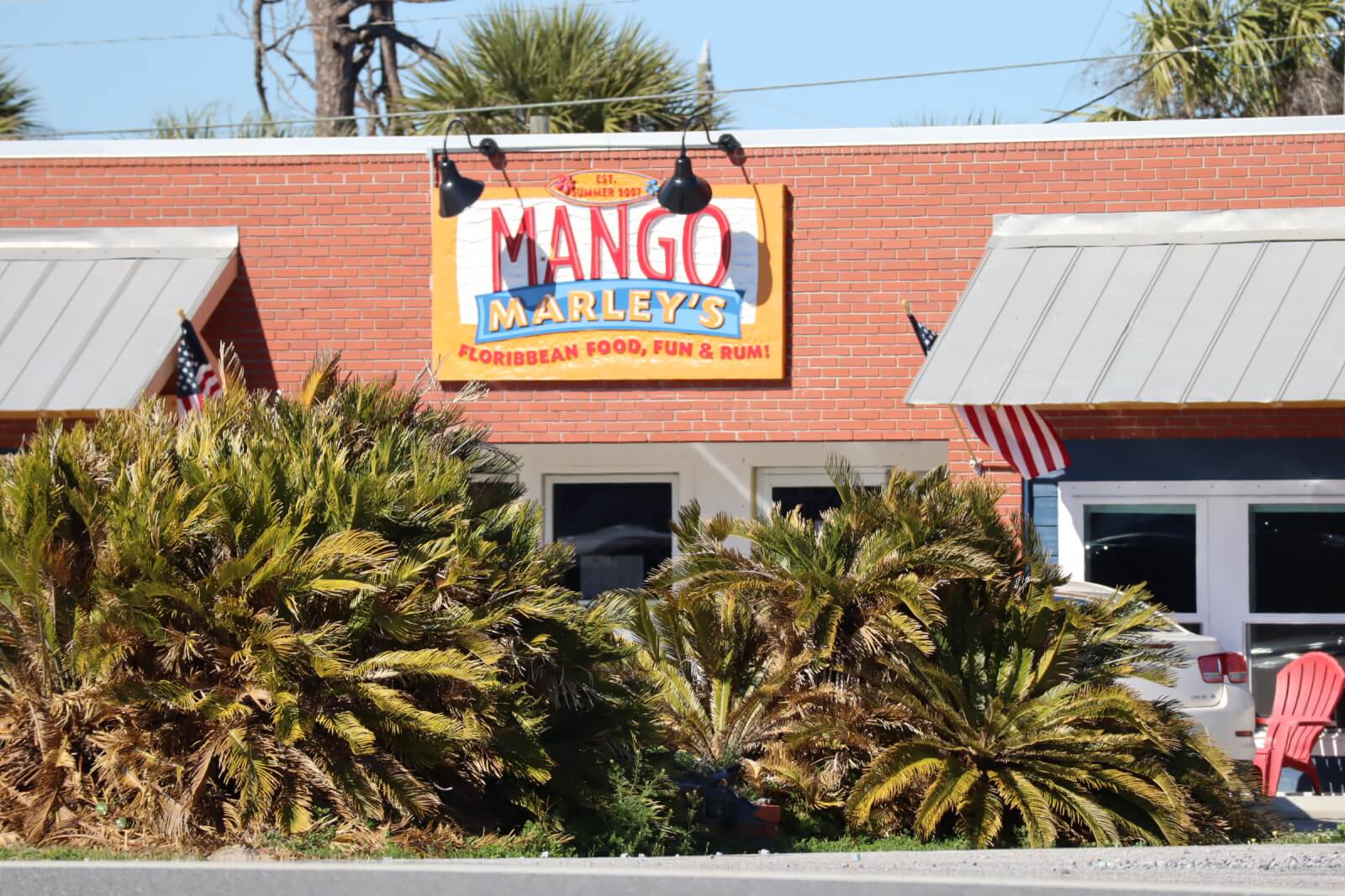 The image captures a rebuilt brick building in a charming Florida town, adorned with a sign that reads "Mango Marley's Caribbean Food, Fun & Rum!" Palm plants and American flags are proudly displayed out front, creating a welcoming post-disaster atmosphere.