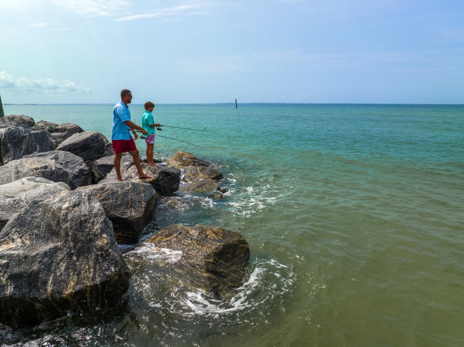 Two people stand on the rocky shore, casting their lines into the ocean's embrace. It's a serene moment in one of Florida's towns, beautifully rebuilt post-disaster, where nature and resilience intertwine seamlessly.