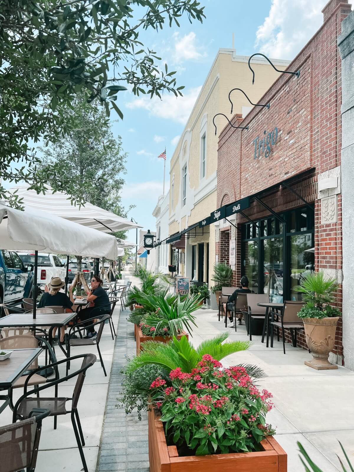 Outdoor seating at a café features tables, chairs, and potted plants along a sidewalk in one of the beautifully rebuilt Florida towns. Diners enjoy their meals as they overlook the charming scene with a brick building sporting a sign reading "Diago" in the background.
