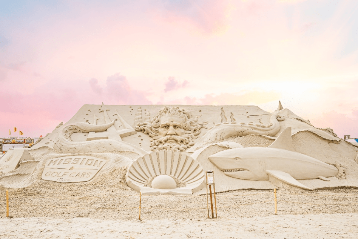 Large sand sculpture featuring a face with flowing hair, ocean waves, a shark, a shell, and a sign reading "MISSION GOLF CARS" under a pastel-colored sky—perfect for celebrating April events by the shore.