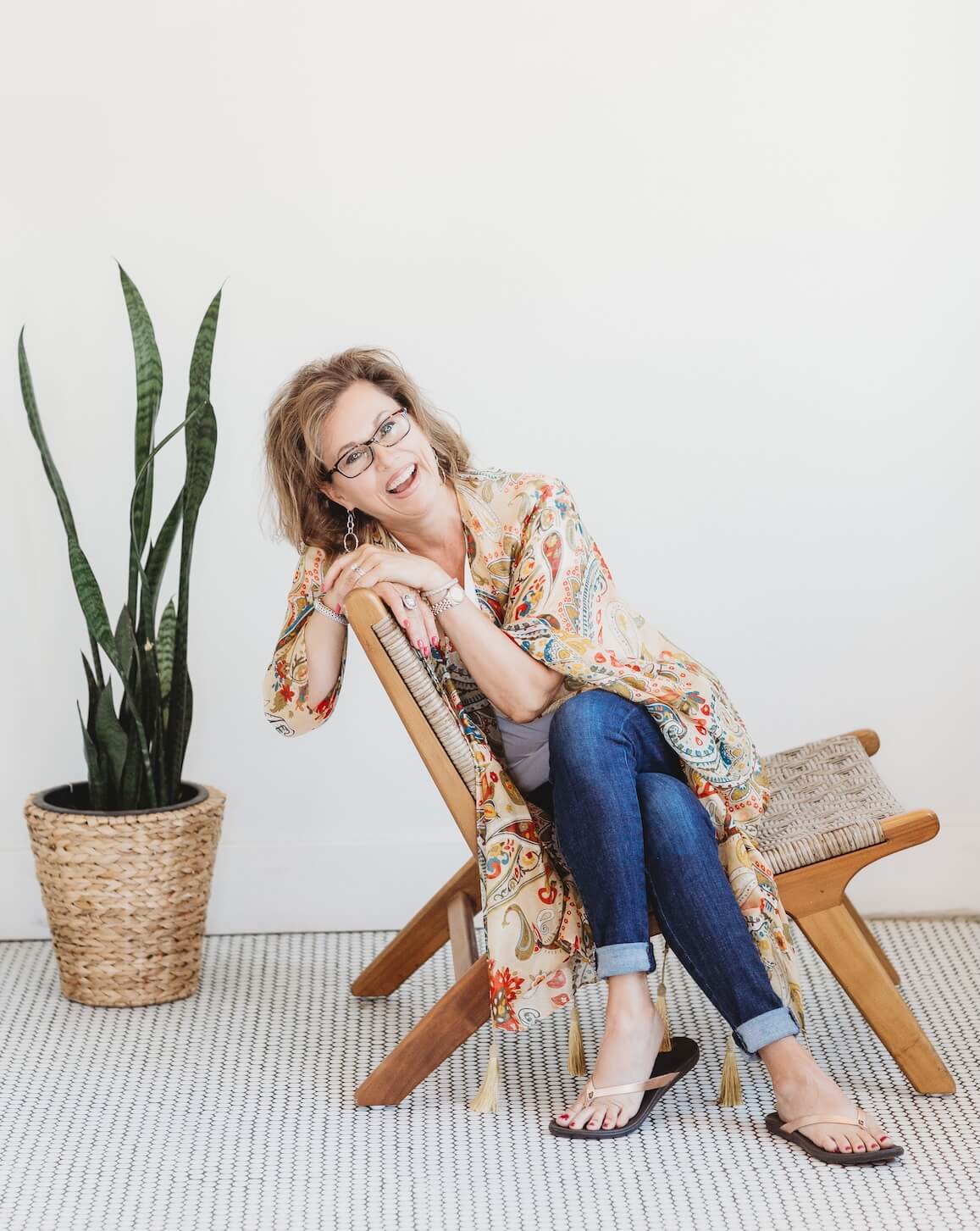 Person seated on a wooden chair, wearing glasses, a floral-patterned cardigan, jeans, and sandals. A potted plant is beside the chair on a light carpet.