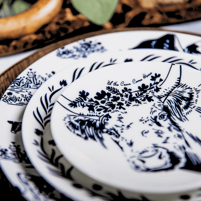 Close-up of stacked plates featuring a black and white cow illustration with floral elements and the phrase "let the cows come home," showcasing the charming artistry of Lovegrass Designs by Kim Jeffers.