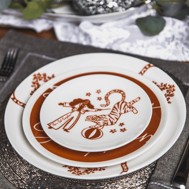 Stacked dinner plates by Lovegrass Designs showcase a circus-themed motif with a tiger and performer, set on a textured mat. Kim Jeffers’ touch is evident, with cutlery, a napkin, and foliage artfully arranged in the background.