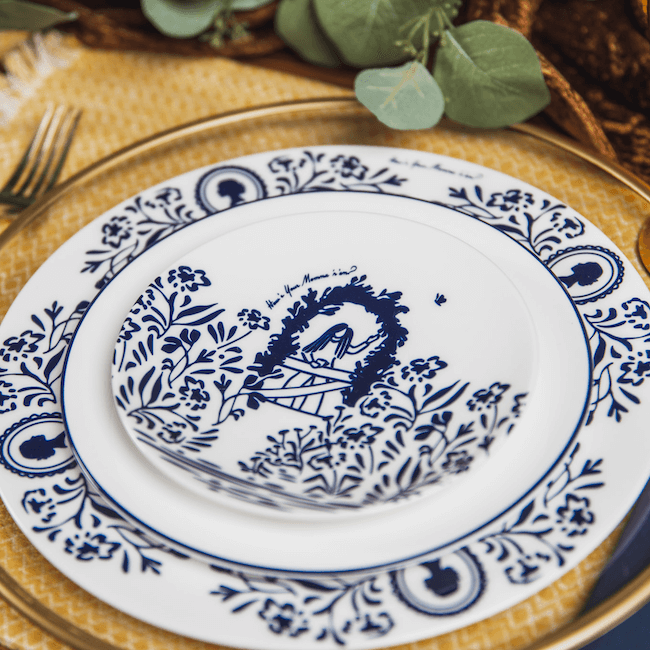 White plates with blue floral patterns from Lovegrass Designs are stacked on a glass charger. They feature a small illustration of a praying figure. A fork and greenery by Kim Jeffers are adjacent on a yellow textured surface.