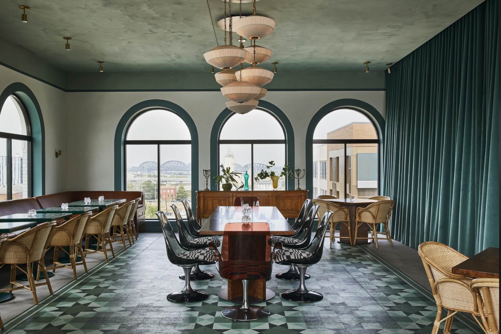 Spacious dining room with patterned floor, long wooden table, black chairs, arched windows, teal curtains, and hanging lamps. 
