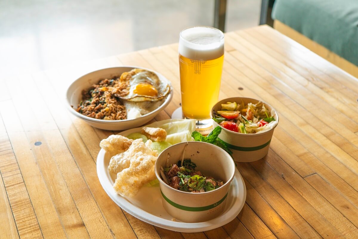Three bowls of assorted Asian dishes, fried egg, rice, vegetables, and pork rinds on a wooden table with a pint of light beer—perfect for exploring flavors at new restaurants in Louisville.