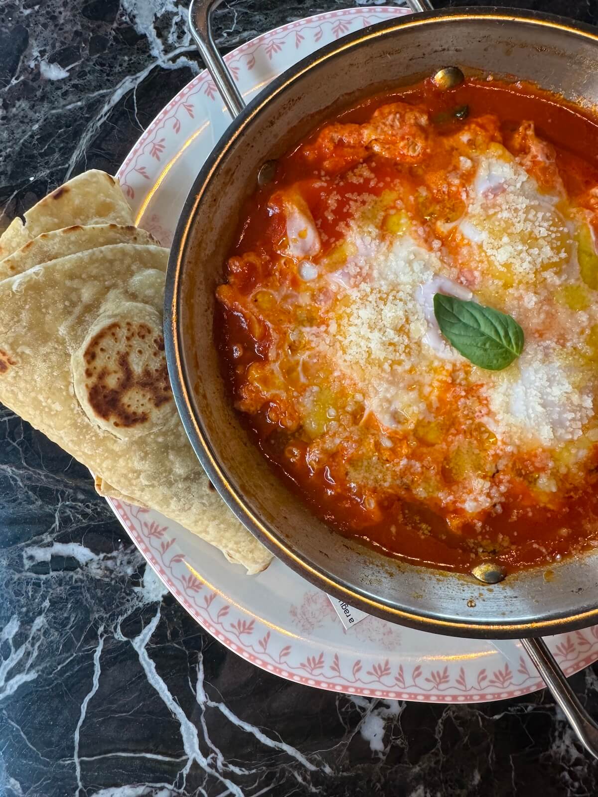 A bowl of tomato-based stew topped with cheese and a basil leaf is served with flatbread on a decorative plate, capturing the flavorful essence found at new Louisville restaurants, and placed on a dark marble table.