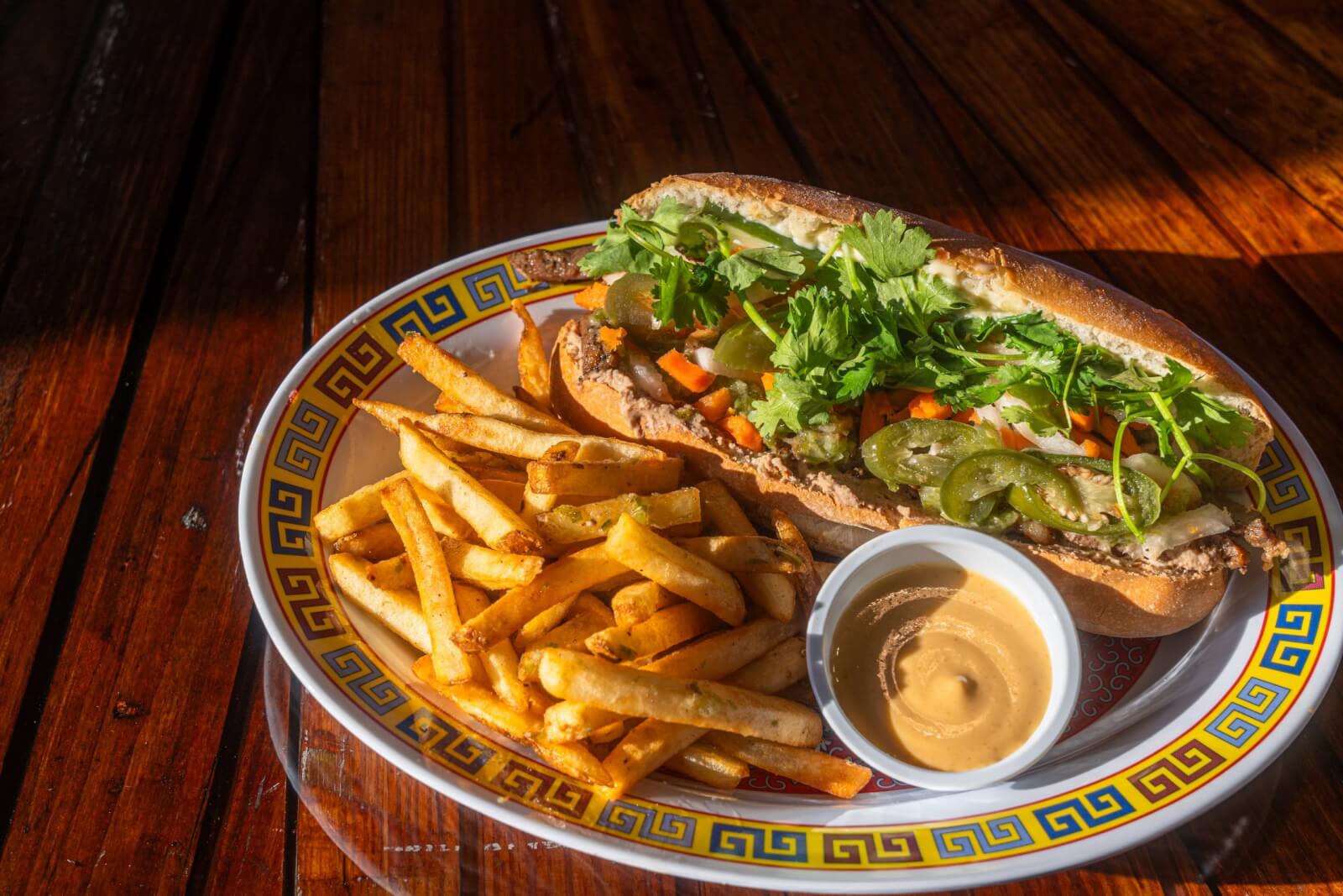 Discover a fresh delight at Louisville's newest restaurants: a vibrant sandwich with crisp vegetables and cilantro on a soft bun, served alongside golden fries and a small cup of sauce, elegantly presented on a decorative plate.