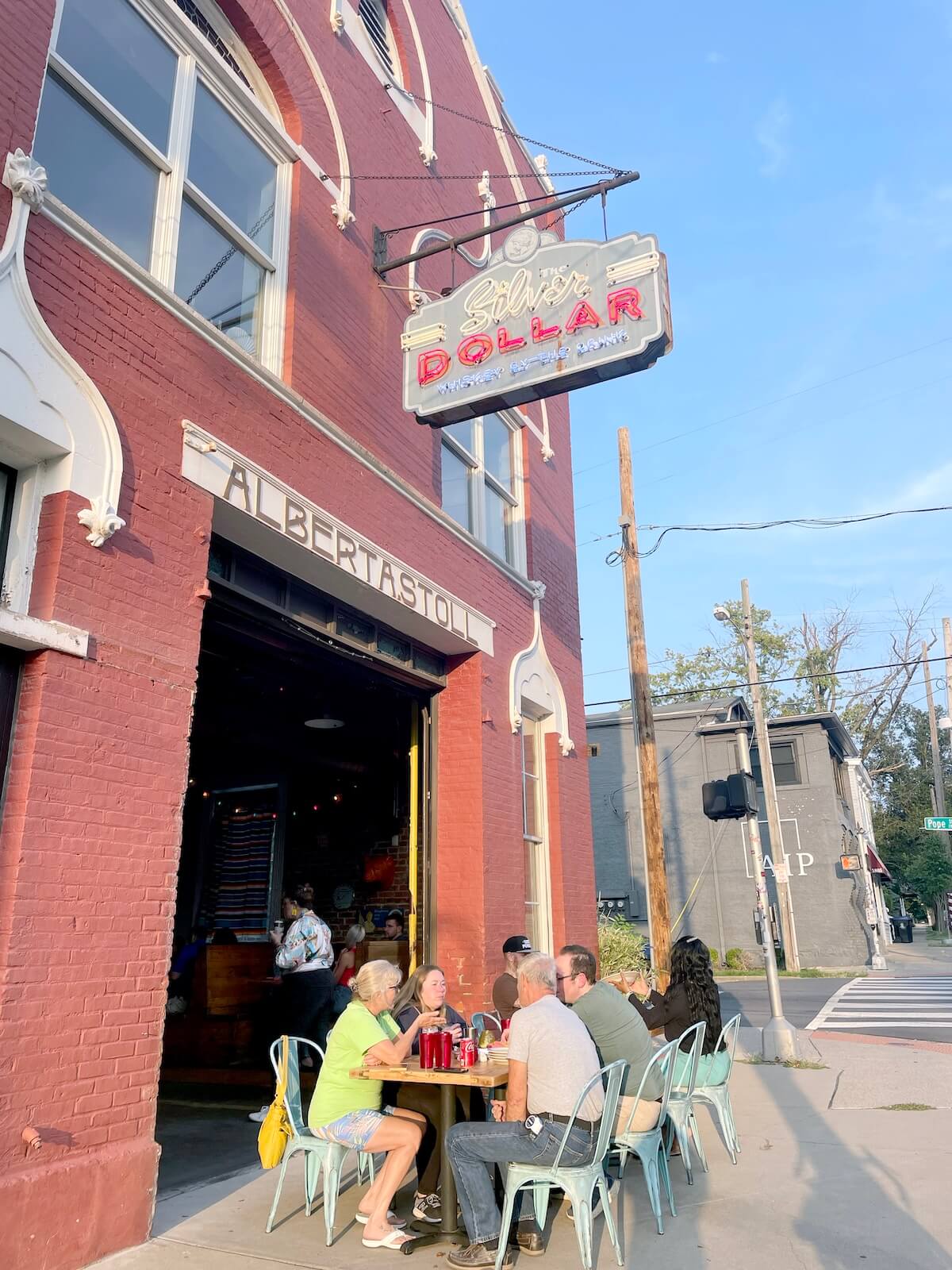 A group of people sit at a table outside a red brick building under a neon “Silver Dollar” sign on a sunny day, enjoying one of the new Louisville restaurants popular for its lively outdoor scene.