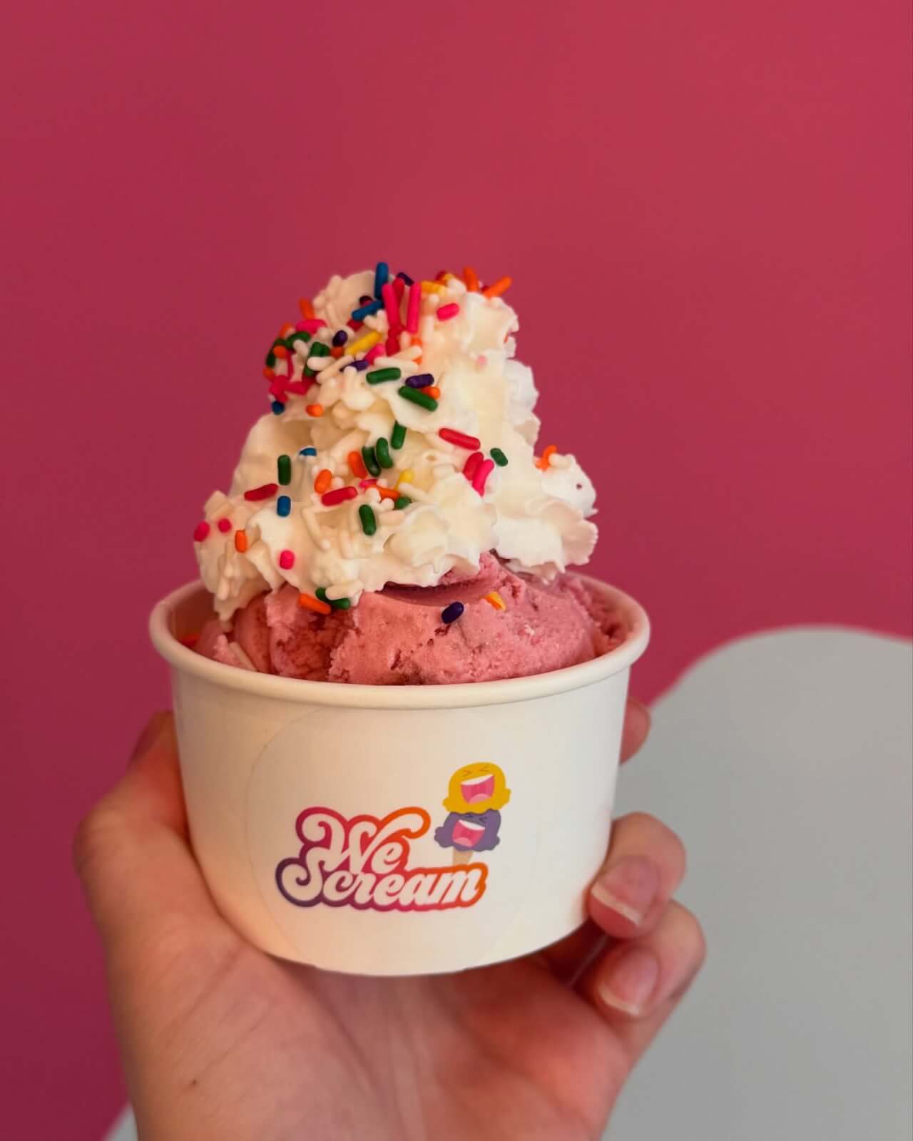 A hand holds a cup of pink ice cream topped with whipped cream and colorful sprinkles, set against a pink backdrop. 