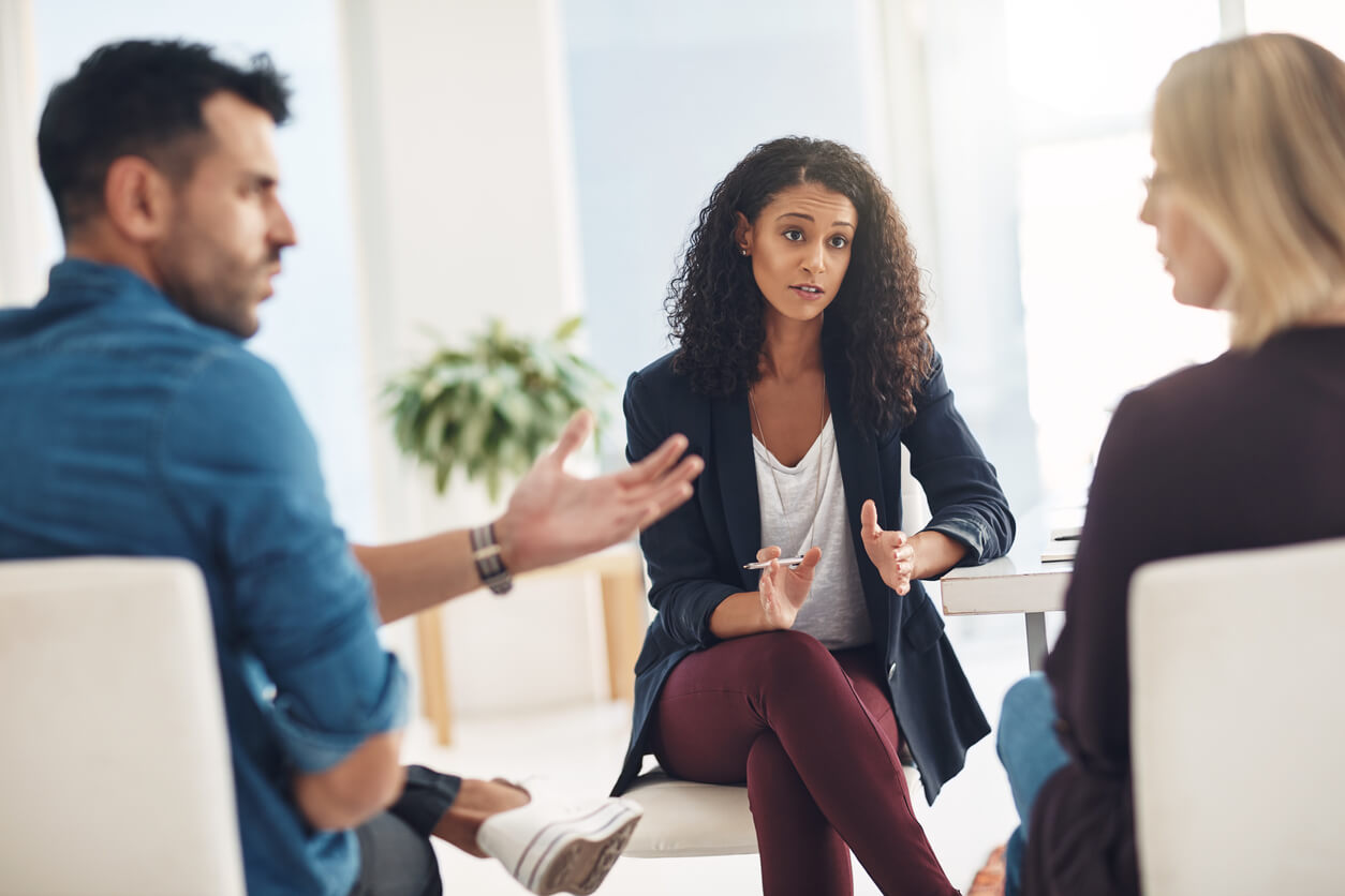 Three people engaged in a serious conversation, sitting indoors, possibly discussing a collaborative divorce process. The woman in the center gestures with her hands while listening to the others. A potted plant is visible in the background, adding a touch of tranquility to their dialogue.