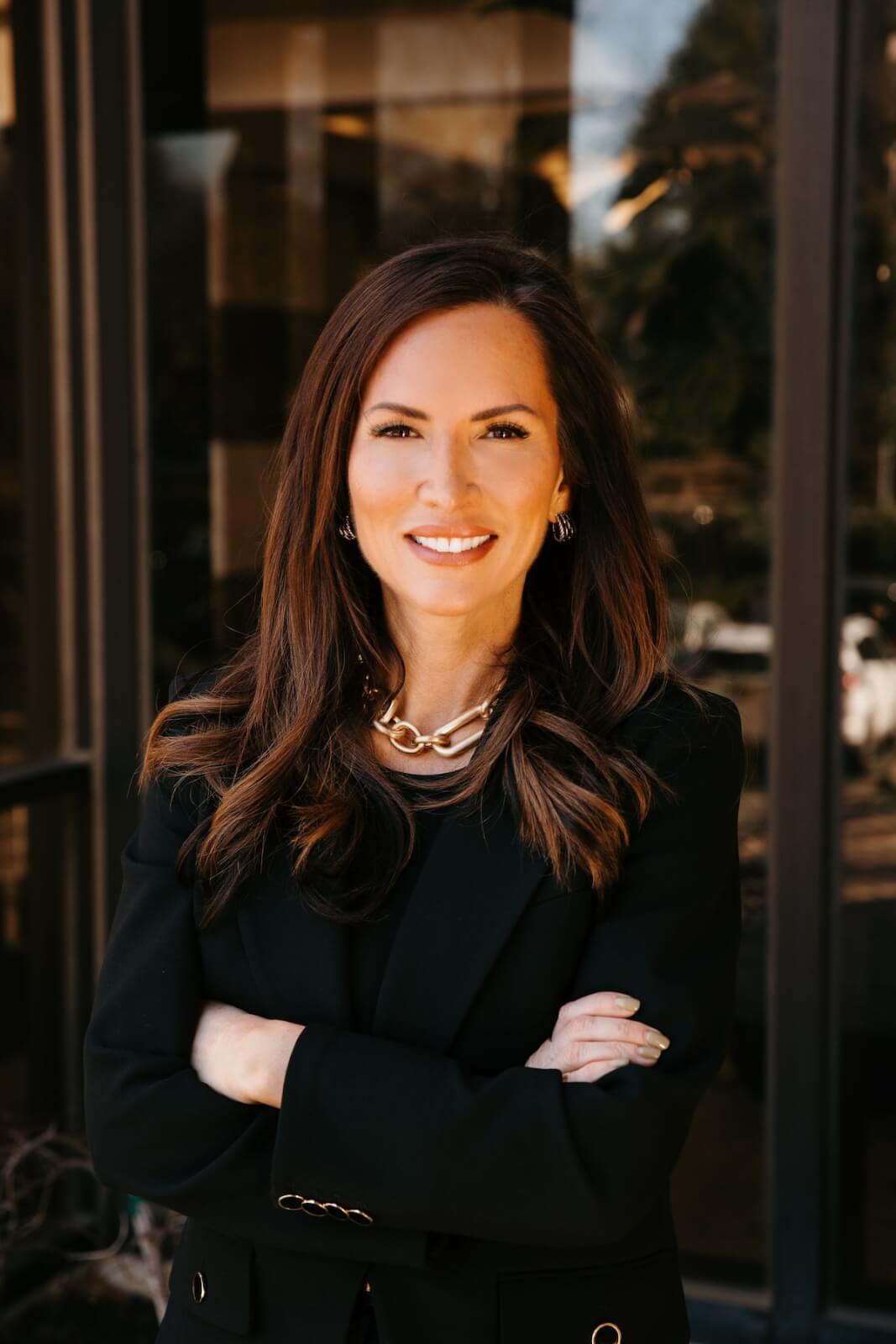 Lisa Gill in a black blazer stands confidently with arms crossed, smiling in front of a glass building.