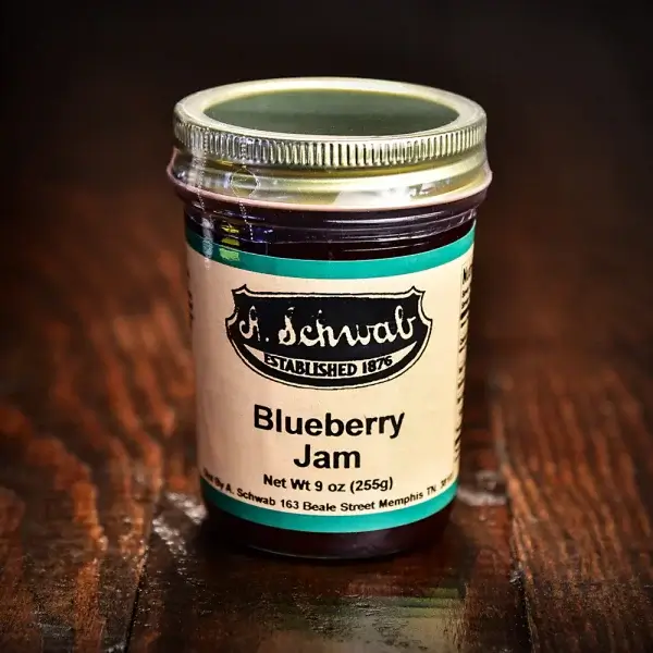 A jar of blueberry jam with a green and beige label, featuring the brand "A. Schwab," established in Memphis in 1876. This April FINDS special is net weight 9 oz (255g) and rests elegantly on a dark wooden surface.
