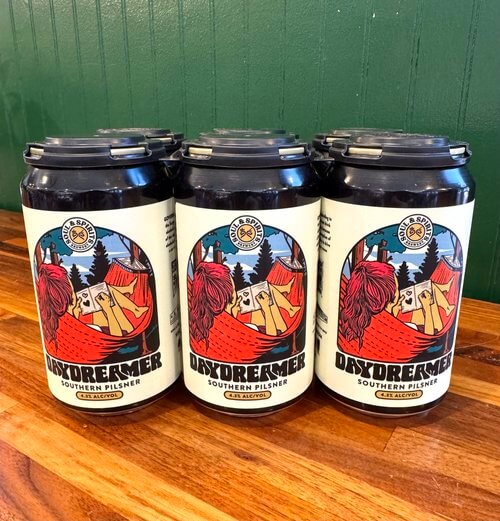 A six-pack of Oxydreamer Southern Pilsner beer rests on a wooden surface, its labels showcasing a colorful outdoor illustration.